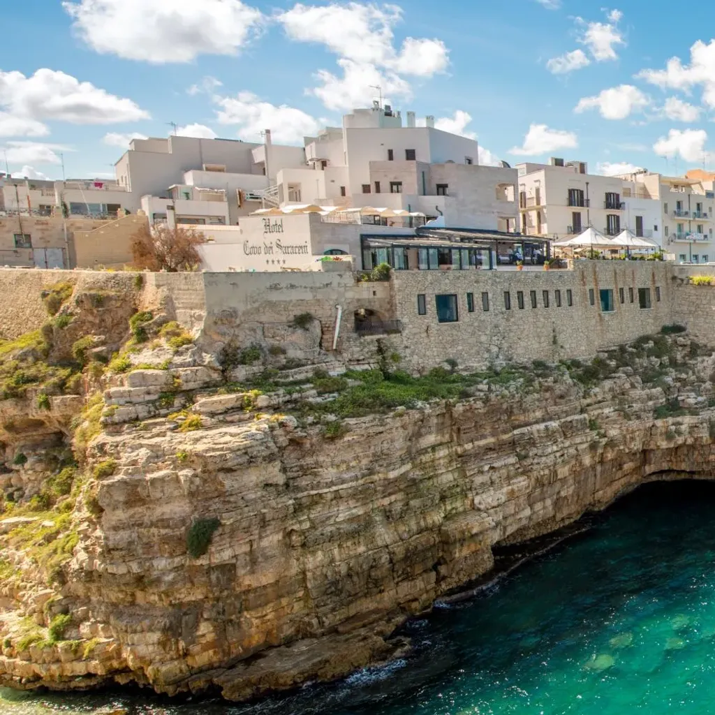 A beautiful panoramic view of a coastal town built atop rugged cliffs, overlooking a stunning turquoise sea under a bright blue sky with scattered white clouds. The ancient buildings with their white and pastel facades contrast beautifully with the vibrant water.