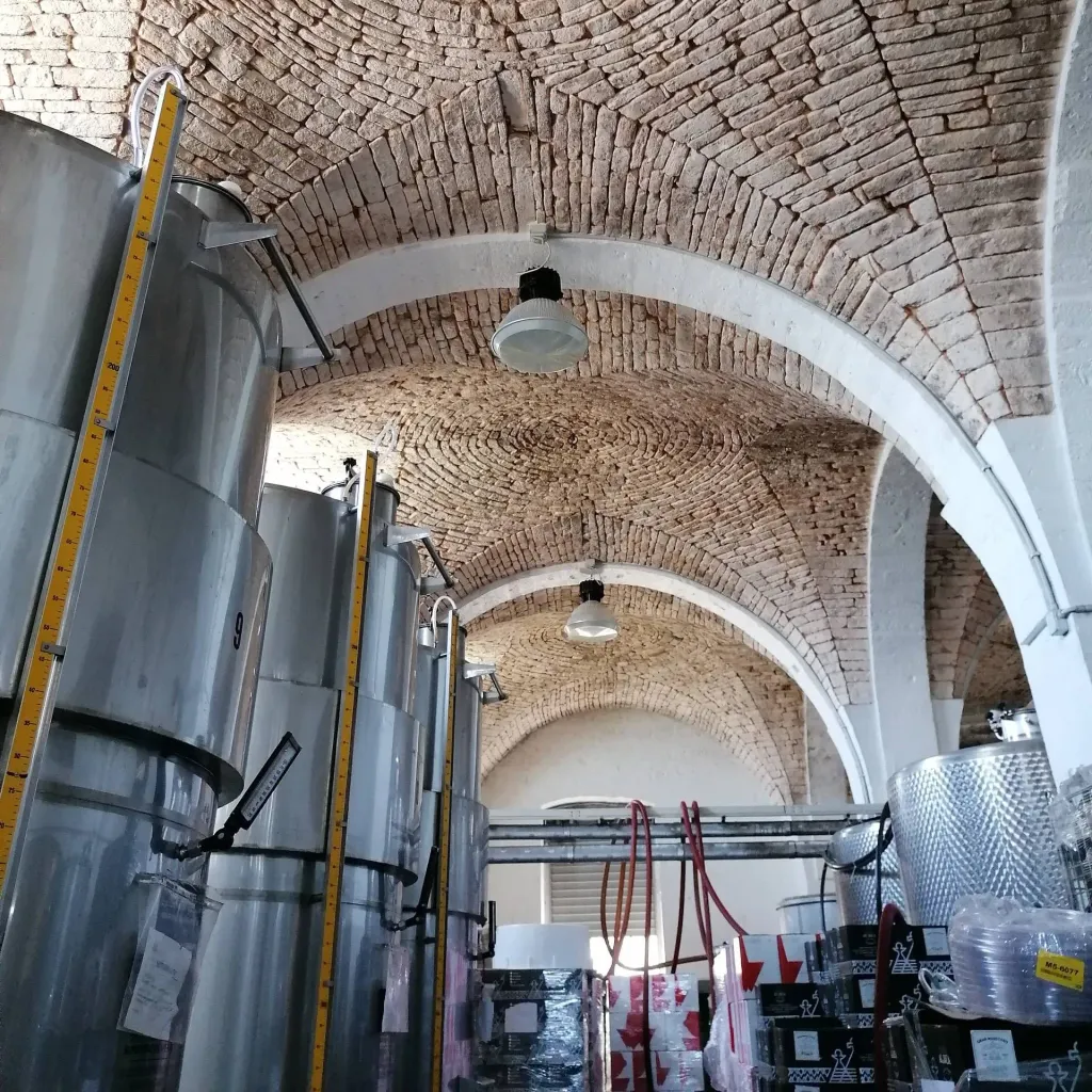 An interior view of a winery, featuring large metal fermentation tanks and an ornate arched brick ceiling.