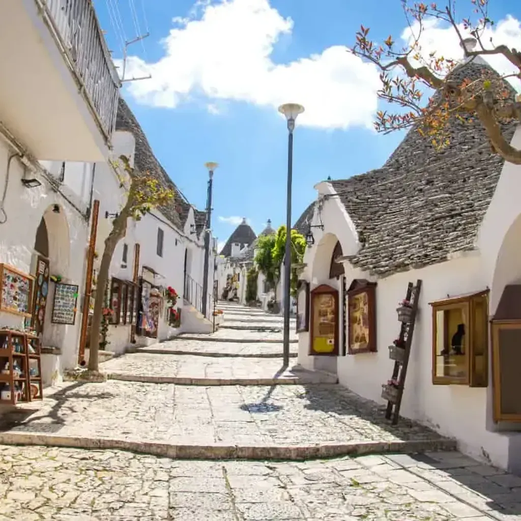 An editorial-style image of a charming, narrow street lined with traditional white-washed houses featuring unique cone-shaped stone roofs, characteristic of Alberobello, Italy. The street is paved with stones and slopes upwards, leading the eye through the scene. The sky is bright blue with some clouds.
